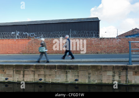 Man and woman walk alongside a canal, with a wall behind topped with razor wire, Loughborough, England. Stock Photo