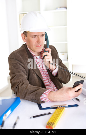 Architect on the phone calculating costs using modern smartphone, working in office at desk. Wearing helmet. Stock Photo