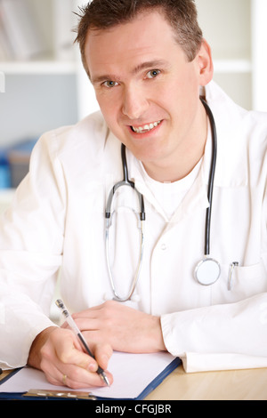 Smiling medical doctor with stethoscope sitting at a desk in his office and taking notes. Stock Photo