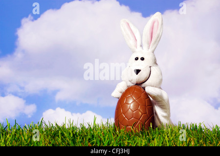 Fun photo of a white easter bunny puppet holding a large chocolate egg on grass with a blue cloudy sky background. Stock Photo