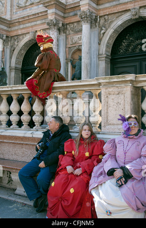 Family in carnival costume sit on a bench looking bored as a carnival character poses on a wall near them St Marks Square Venice Stock Photo