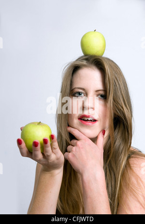Attractive young Caucasian woman having fun with apples with one in hand and one balancing on head Stock Photo