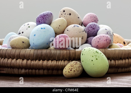 Still life photo of speckled candy covered chocolate easter eggs in a wicker basket on a rustic wooden table.