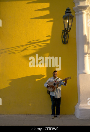 A male Hispanic street musician plays his guitar against a yellow wall late afternoon with strong shadows in Havana, Cuba. Stock Photo