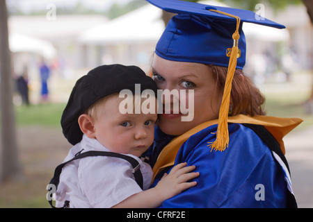 A young mother in graduation gown and cap posing with her small boy. The boy is wearing suspenders and cap. Stock Photo