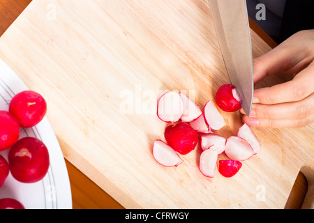 Detail of chopping board and female hand with knife cutting fresh radishes. Stock Photo