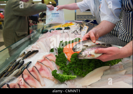 A fish monger holding two rainbow trout at the fish counter in a UK supermarket while a customer is served in the background. Stock Photo