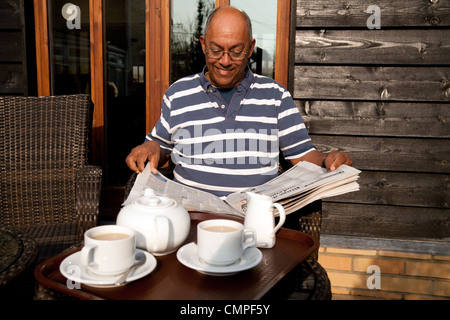 A middle aged Indian man reading the daily newspaper while having a cup of tea, Cambridgeshire, UK Stock Photo