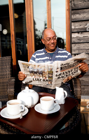 A middle aged Indian man reading the newspaper while having a cup of tea, Cambridgeshire, UK Stock Photo