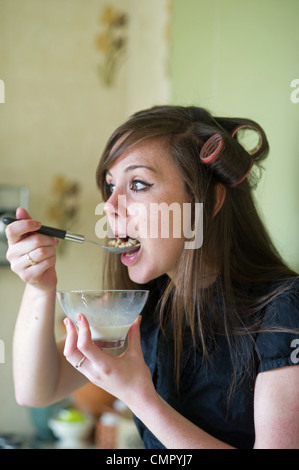 Young woman in twenties 20s age group wearing hair rollers eating breakfast in rush to get to work UK - posed by model Stock Photo