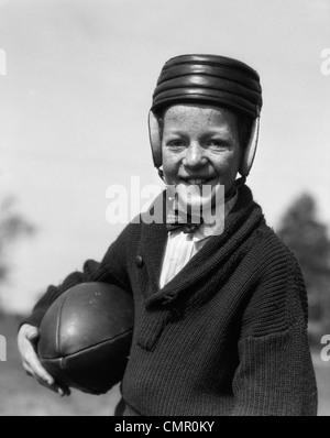 1920s FRECKLE-FACED BOY IN SWEATER & BOW TIE WEARING LEATHER FOOTBALL HELMET HOLDING FOOTBALL UNDER ARM LOOKING AT CAMERA Stock Photo