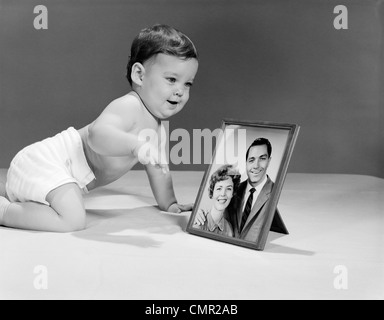 1960s BABY IN DIAPER CRAWLING TO FRAMED PICTURE OF MOTHER AND FATHER Stock Photo