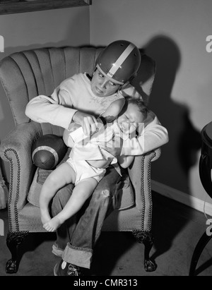 1950s 1960s BOY SITTING BY FOOTBALL IN EASY CHAIR WEARING HELMET TRYING TO HOLD AND FEED BOTTLE TO CRYING BABY Stock Photo