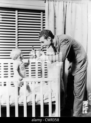 1930s 1940s BABY STANDING IN CRIB IN FRONT OF WINDOW WITH VENETIAN BLINDS FATHER HANDING BABY A TOY DOG STUFFED ANIMAL NURSERY Stock Photo