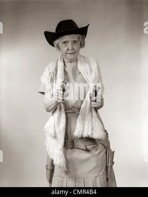 1950s GRANNY COWGIRL WEARING HAT & SHAWL & POINTING 2 PISTOLS AND LOOKING AT CAMERA Stock Photo
