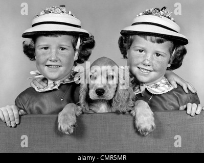 1950s 1960s TWIN GIRLS WEARING WHITE STRAW HATS SEPARATED BY A COCKER SPANIEL PUPPY LOOKING OVER A STUDIO FENCE INSIDE Stock Photo