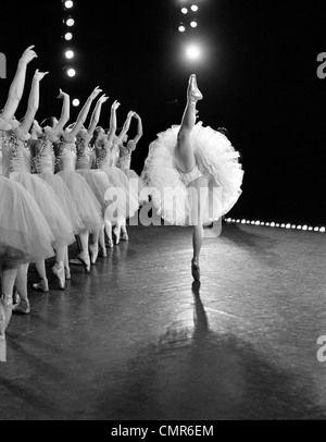 1970s SIDE VIEW OF PRIMA BALLERINA IN STANDING SPLIT WITH ROW OF 8 BACKING DANCERS WITH ARMS RAISED BEHIND HER Stock Photo