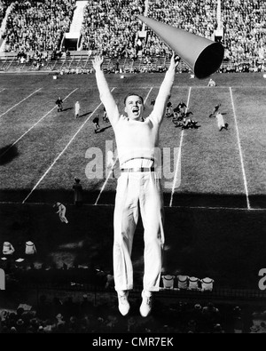 1940s MALE CHEERLEADER WITH MEGAPHONE JUMPING IN AIR AT FOOTBALL GAME Stock Photo