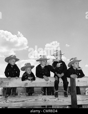 1950s FIVE BOYS BROTHERS DRESSED AS COWBOYS SITTING TOGETHER ON CORRAL FENCE Stock Photo