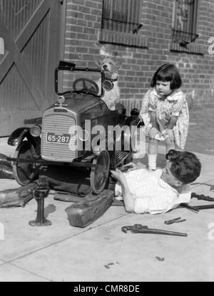 1920s LITTLE GIRL LEANING DOWN WATCHING BOY WORKING UNDER TOY CAR WITH DOG BEHIND WHEEL Stock Photo