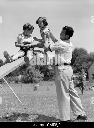 1950s FATHER LIFTING SON AND DAUGHTER ONTO A PLAYGROUND SEESAW OUTDOOR Stock Photo