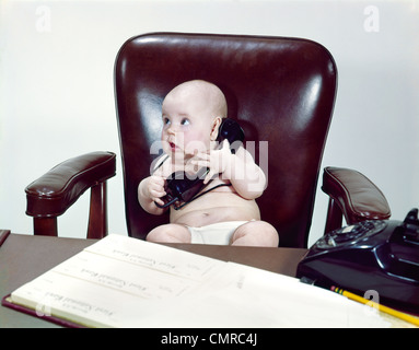 1960s CHUBBY BABY SITTING IN LEATHER CHAIR AT OFFICE DESK HOLDING TELEPHONE Stock Photo