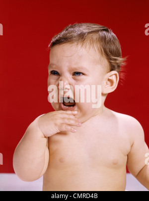 1960s PORTRAIT OF CRYING BABY HOLDING HAND UP TO MOUTH RED BACKGROUND Stock Photo