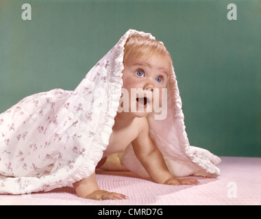 1960s BABY CRAWLING AND PEEKING OUT FROM UNDER A BLANKET Stock Photo