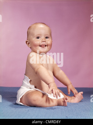 1960s SMILING BABY WEARING CLOTH DIAPER SITTING ON BLUE BLANKET STICKING OUT TONGUE LOOKING AT CAMERA Stock Photo