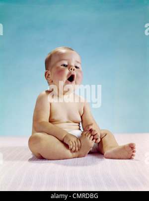 1960s TIRED BABY SITTING HOLDING ON TO ONE FOOT YAWNING FACIAL EXPRESSION Stock Photo