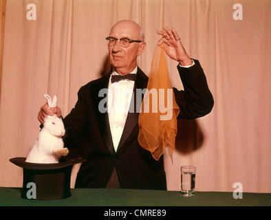 1960s 1970s MAGICIAN PULLING A WHITE RABBIT OUT OF HAT AND HOLDING AN ORANGE SCARF Stock Photo