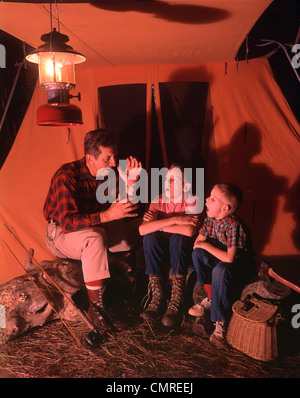 1960s 1970s  GRANDFATHER TELLING SCARY STORY TO BOYS BY TENT AT NIGHT CAMPSITE IN SHADOWS Stock Photo