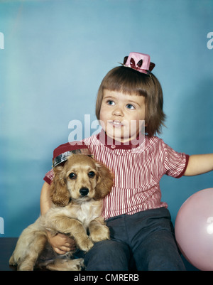 1950s 1960s LITTLE GIRL IN PARTY HAT SITTING HOLDING A COCKER SPANIEL PUPPY Stock Photo