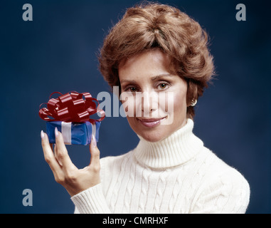 1970s PORTRAIT OF SMILING WOMAN WEARING CABLE KNIT TURTLENECK SWEATER HOLDING SMALL WRAPPED GIFT LOOKING AT CAMERA Stock Photo