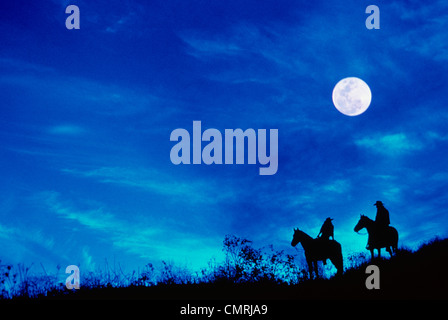 1980s SILHOUETTE OF TWO COWBOYS ON HORSES AT TWILIGHT FULL MOON IN SKY Stock Photo