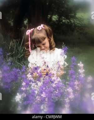 1970s SMILING LITTLE GIRL IN WHITE DRESS PINK HAIR RIBBONS LOOKING AT PURPLE FLOWERS Stock Photo