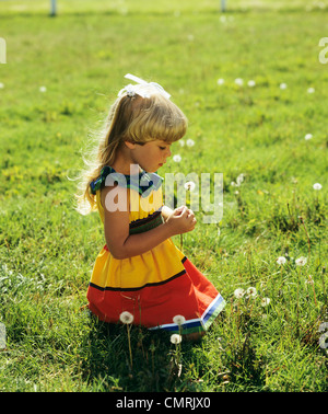 1980s LITTLE BLOND GIRL WEARING RED AND YELLOW SUN DRESS KNEELING ON GRASS LOOKING AT DANDY LION FLOWER SEED HEAD Stock Photo