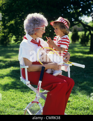 1980s GRANDMOTHER SITTING IN DECK CHAIR HOLDING TENNIS RACQUET AND GRANDDAUGHTER WEARING TENNIS CLOTHES Stock Photo
