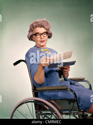 1960s SENIOR WOMAN WEARING GLASSES SITTING IN WHEELCHAIR READING LETTER INSURANCE CHECK HEALTHCARE PATIENT Stock Photo