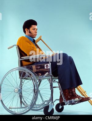 1970 1970s RETRO AFRICAN AMERICAN MAN SITTING IN WHEELCHAIR WITH CRUTCHES SAD SERIOUS EXPRESSION PATIENT HEALTHCARE Stock Photo