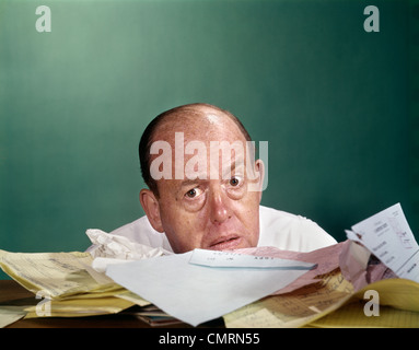 1960s BALDING MIDDLE AGED MAN WORRIED EXPRESSION PEEKING OVER TOP PAPERS BILLS PAPERWORK Stock Photo