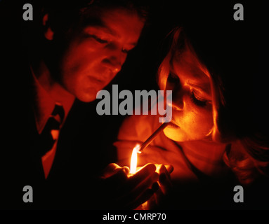 1970s CLOSE UP OF WOMAN LIGHTING CIGARETTE FROM FLAME MAN IS HOLDING TOBACCO NOSTALGIA HEALTH HAND Stock Photo