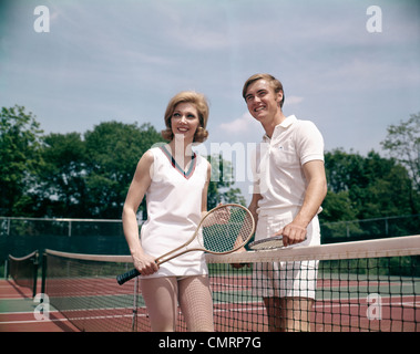 1970 1970s RETRO SMILING COUPLE MAN WOMAN STANDING ON OPPOSITE SIDES OF NET TENNIS COURT HOLDING RACKETS RECREATION Stock Photo