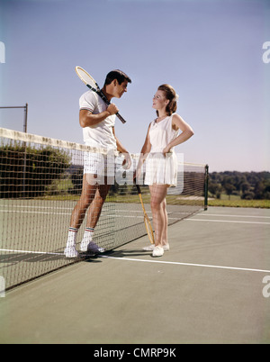 1960 1960s RETRO SMILING COUPLE MAN WOMAN STANDING ON OPPOSITE SIDES OF NET TENNIS COURT HOLDING RACKETS RECREATION Stock Photo