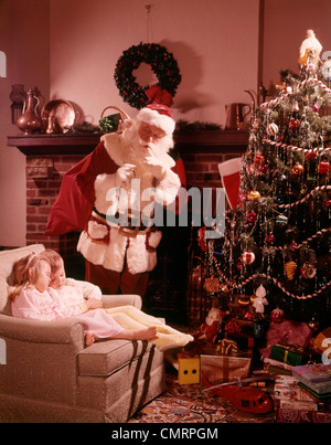 1960 1960s TWO CHILDREN BOY GIRL ASLEEP CHAIR BY FIREPLACE CHRISTMAS TREE SANTA LOOKING DOWN BY TREE WITH PRESENTS RETRO Stock Photo