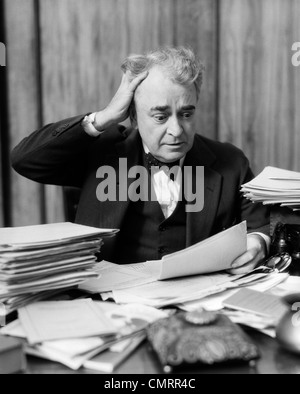 1930s ELDERLY BUSINESSMAN SITTING AT DESK AMONG STACKS OF PAPERS WITH HAND IN HAIR & FLUSTERED EXPRESSION Stock Photo
