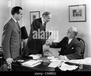 1930s 1940s TWO SALESMEN MAKING SALE TO BUSINESSMAN SITTING AT DESK HAND SHAKE TO CLOSE THE DEAL SMILING Stock Photo