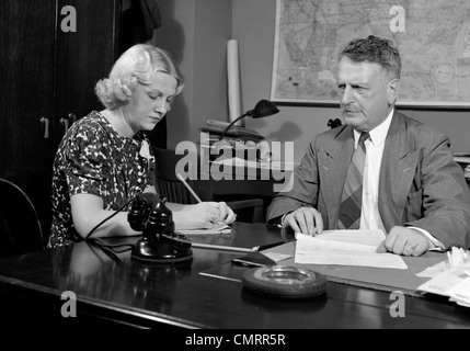 1930s 1940s BUSINESSMAN SITTING AT DESK GOING OVER PAPERWORK WITH SECRETARY TAKING DICTATION MAN WOMAN OFFICE INDOOR Stock Photo
