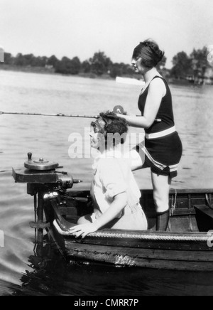 1920s 1930s TWO WOMEN FISHING FROM STERN OF SMALL BOAT WITH MOTOR OUTDOOR ONE WOMAN WI WEARING BATHING SUIT Stock Photo