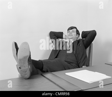 1970s CONFIDENT YOUNG BUSINESSMAN SITTING RELAXED WITH FEET UP ON DESK Stock Photo